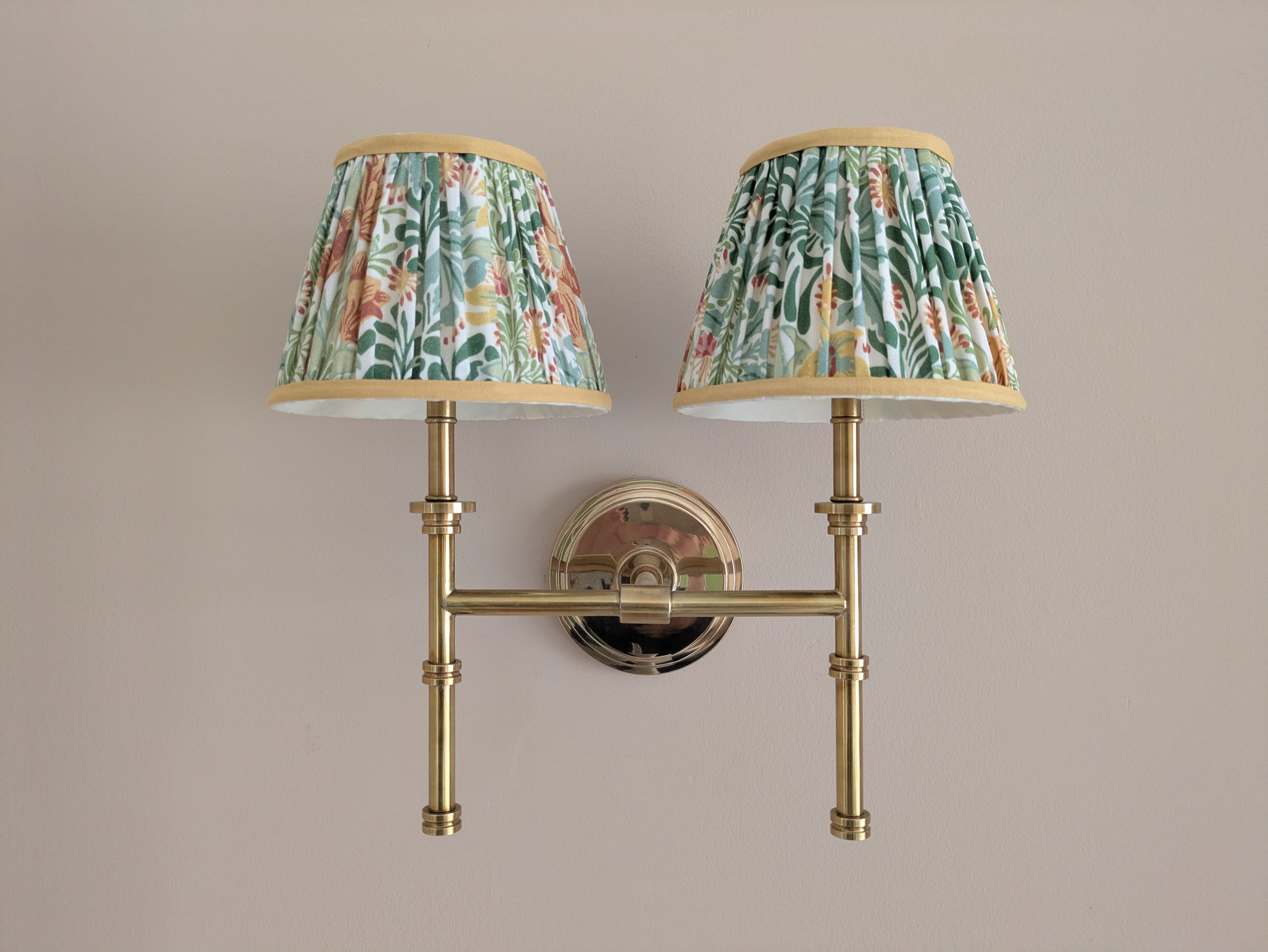 A brass double wall lamp with two pleated lampshades in a green and pink floral print.