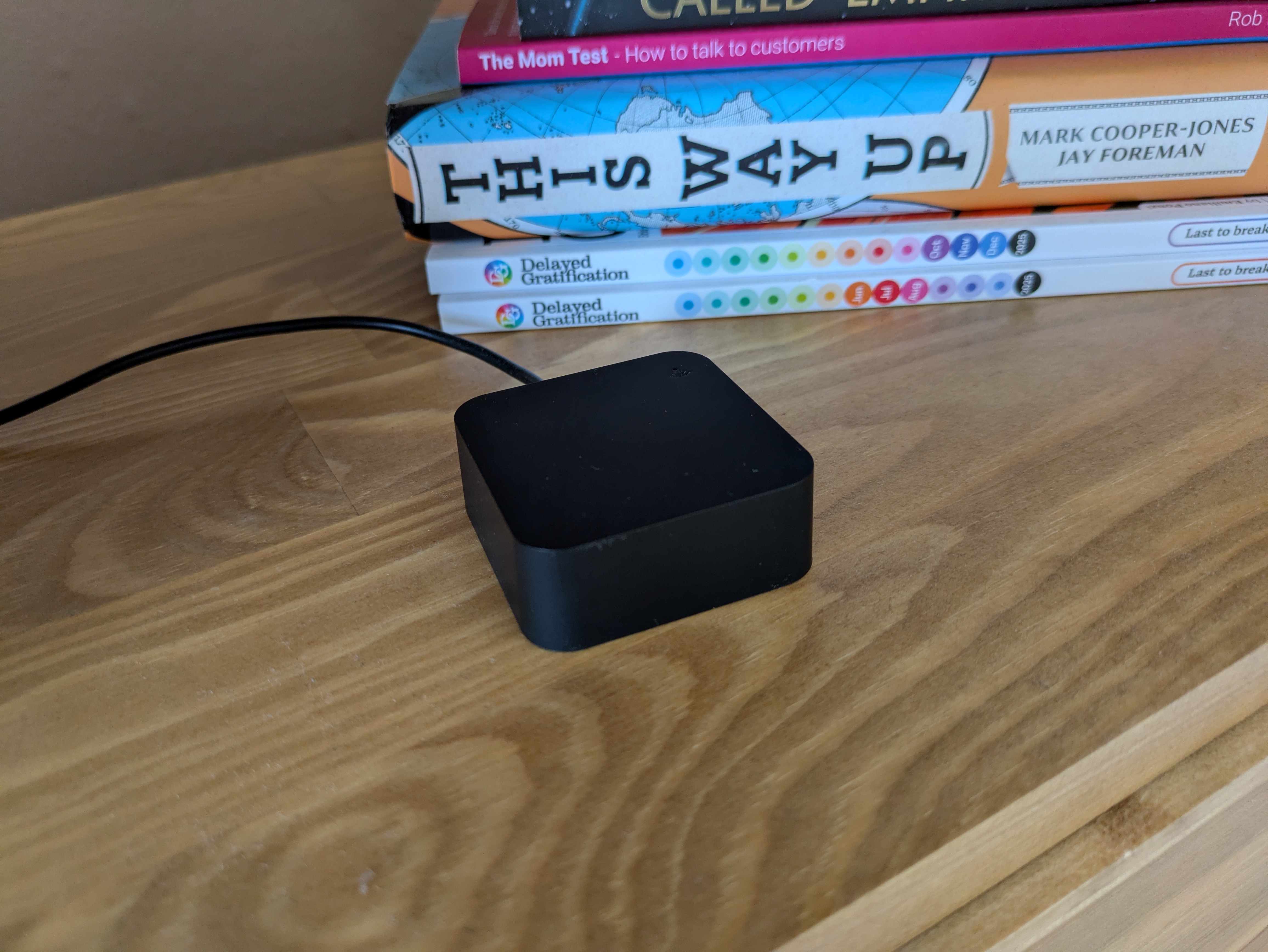 A small, featureless black plastic cube with a cable trailing from it, sitting on a wooden shelf in front of a stack of books.